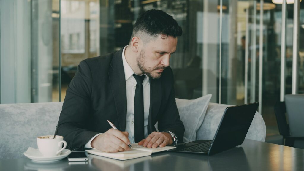 Geschäftsmann im Anzug notiert im Café Ideen in ein Notizbuch neben dem Laptop – Vorbereitung für LinkedIn Personal Branding