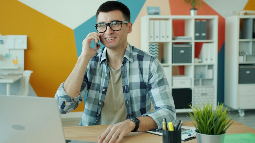 Lächelnder Mitarbeiter mit Brille telefoniert am Schreibtisch, um beim Rebranding planen wichtige Kunden persönlich zu informieren